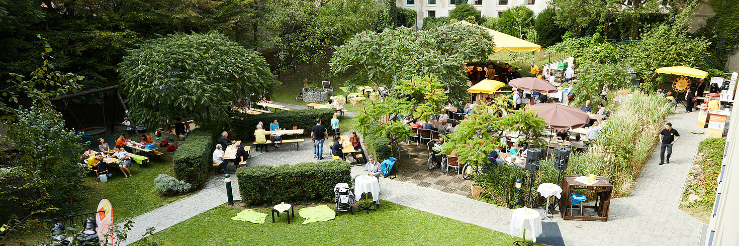Mehrere Menschen feiern in einem großen Garten ein sommerliches Fest. Es gibt eine kleine Bühne und die Leute sitzen gemeinsam an Tischen.