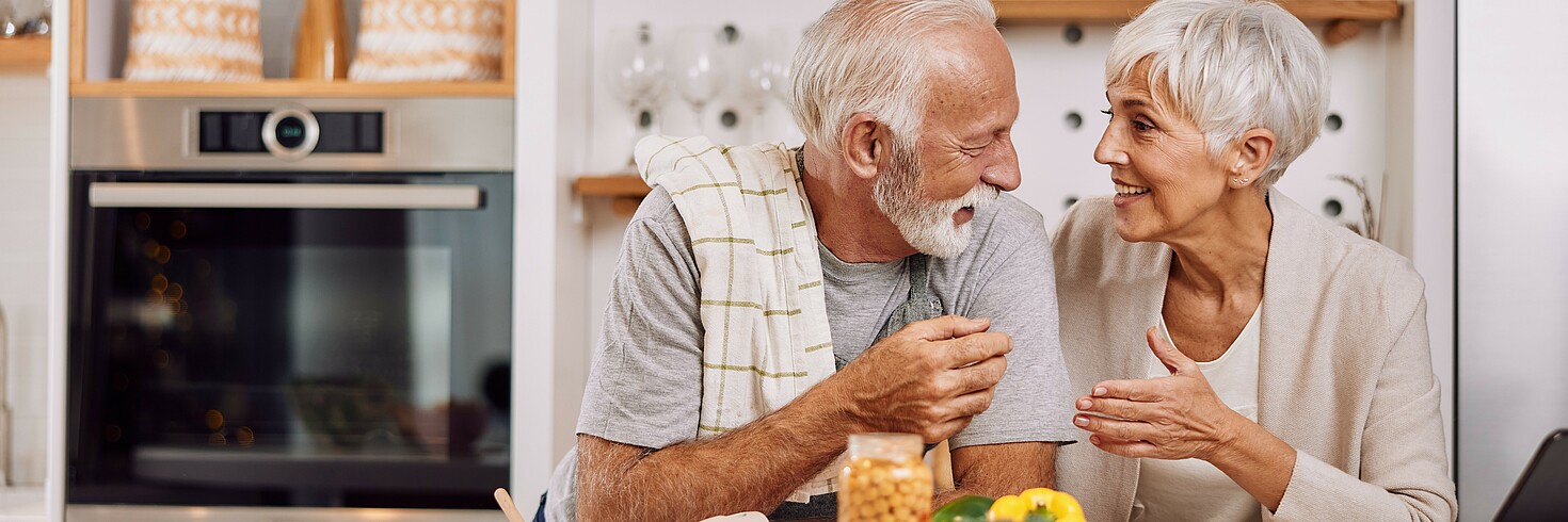 Ein älteres Paar steht in einer Küche. Im Vordergrund liegen gesunde Zutaten. Das Paar lächelt sich an.
