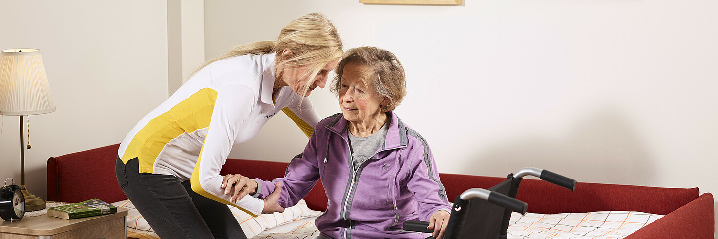 Hilfswerk-Mitarbeiterin hilft älterer Dame aus dem Bett in ihren Rollstuhl.