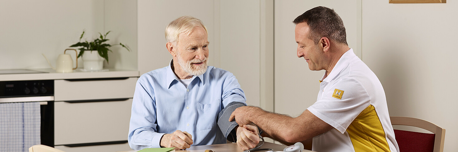 Hilfswerk-Mitarbeiter sitzt mit älterem Herrn an einem Tisch und misst seinen Blutdruck.
