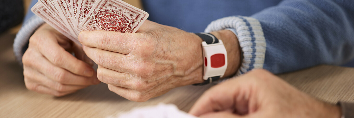 Zwei ältere Damen und zwei ältere Herren spielen Karten, einer der Herren besitzt ein Notrufarmband.