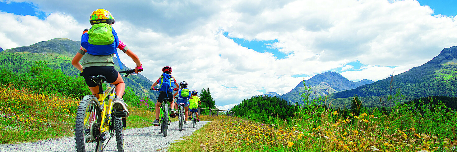 Mehrere Kinder fahren hintereinander auf Fahrrädern einen Weg entlang, rundherum sieht man grüne Wiesen und Berge