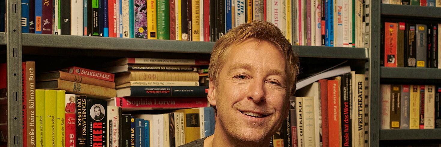Bild von einem Mann mit blonden kurzen Haaren vor einem Bücherregal