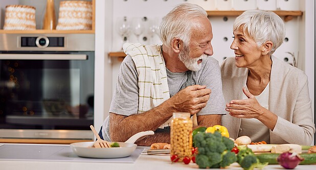Ein älteres Paar steht in einer Küche. Im Vordergrund liegen gesunde Zutaten. Das Paar lächelt sich an.