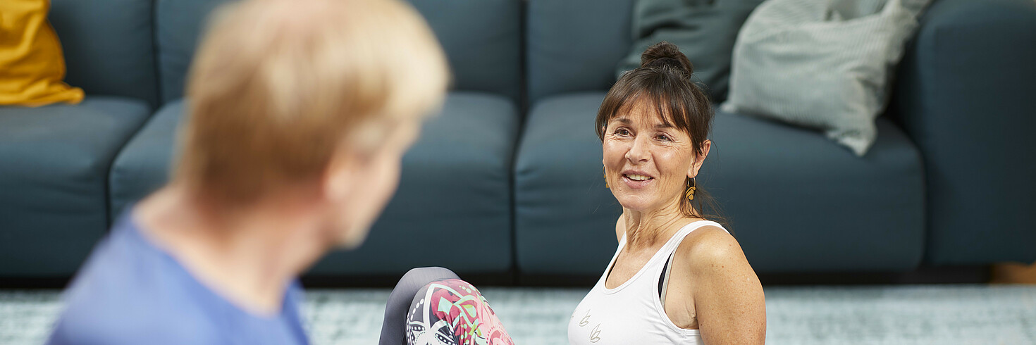 Ein Paar mittleren Alters sitzt im Wohnzimmer am Boden. Die Frau sitzt auf einer Gymnastikmatte und stützt sich auf, der Mann sitzt mit dem Rücken zur Kamera. Sie schauen sich an. 