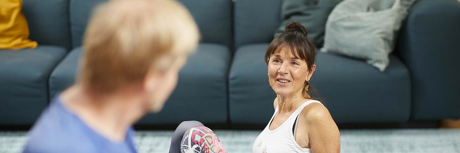 Ein Paar mittleren Alters sitzt im Wohnzimmer am Boden. Die Frau sitzt auf einer Gymnastikmatte und stützt sich auf, der Mann sitzt mit dem Rücken zur Kamera. Sie schauen sich an. 