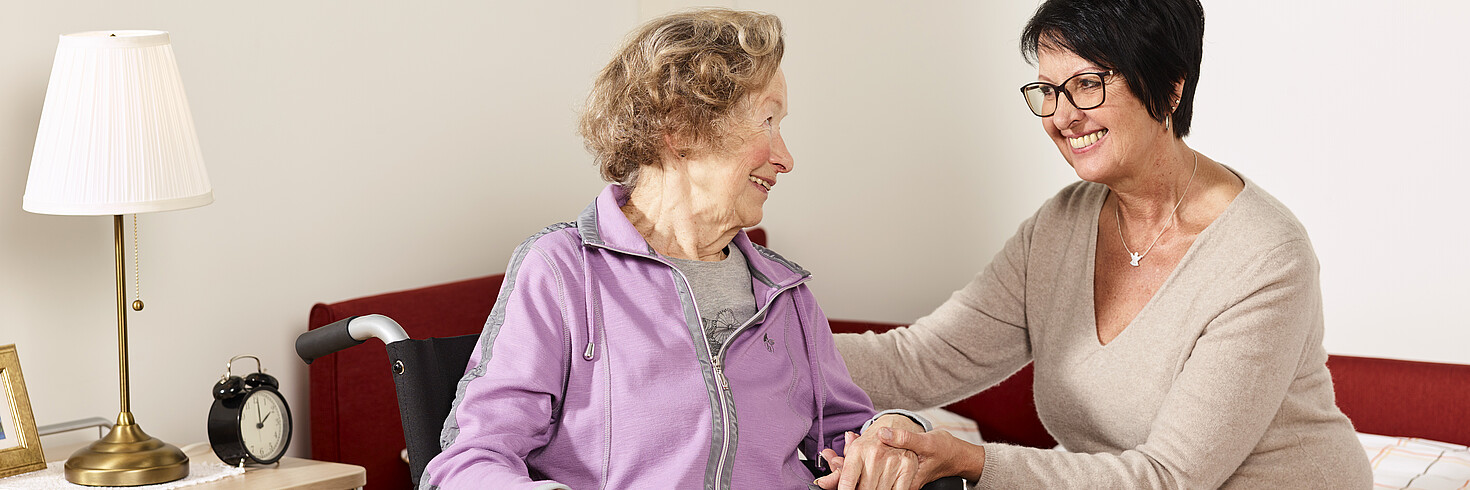 Jüngere Frau hält älterer Dame im Rollstuhl die Hand, beide lächeln