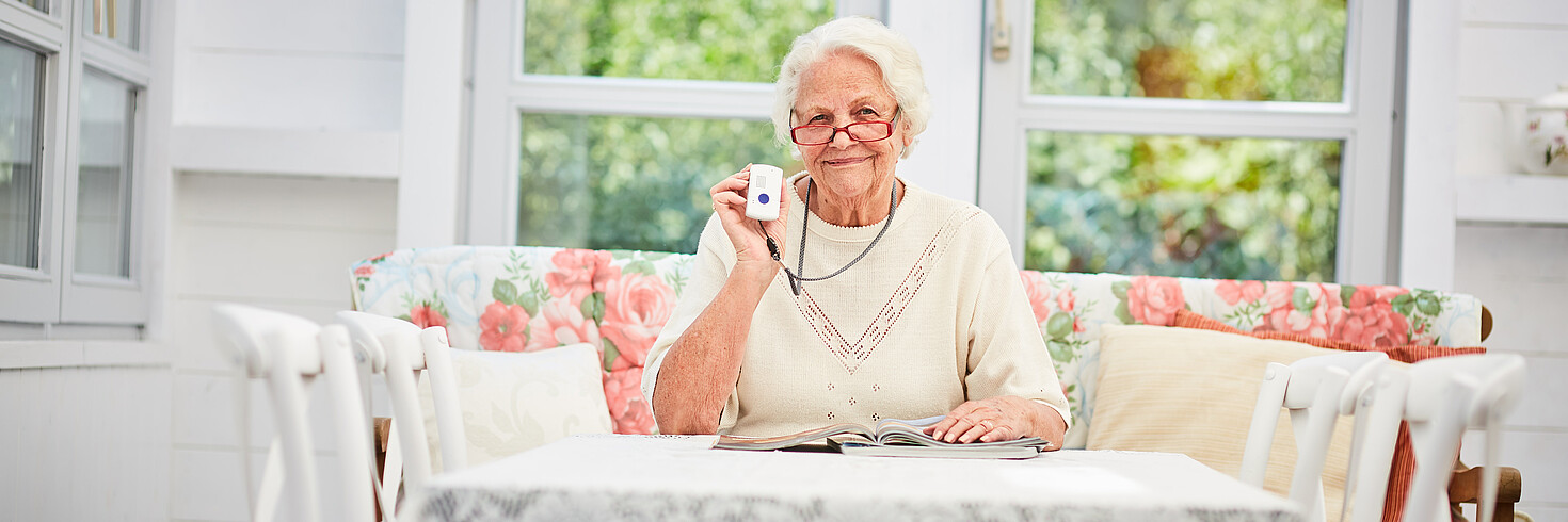 Ältere Dame mit Notruftelefon sitzt lesend in ihrem Wohnzimmer.