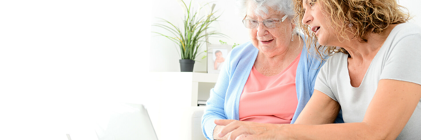 Ältere Dame sitzt mit 24-Stunden-Betreuerin am Sofa vor dem Laptop