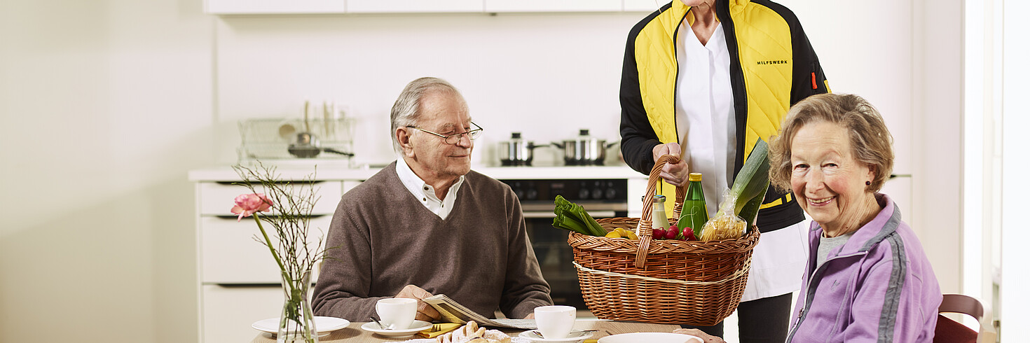 Älteres Ehepaar sitzt an einem Tisch, hinter ihnen steht eine Hilfswerk-Mitarbeiterin, die Einkäufe in der Hand.