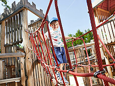 Bub im Vorschulalter auf einem Klettergestell aus Holz und Seilen lächelt in die Kamera