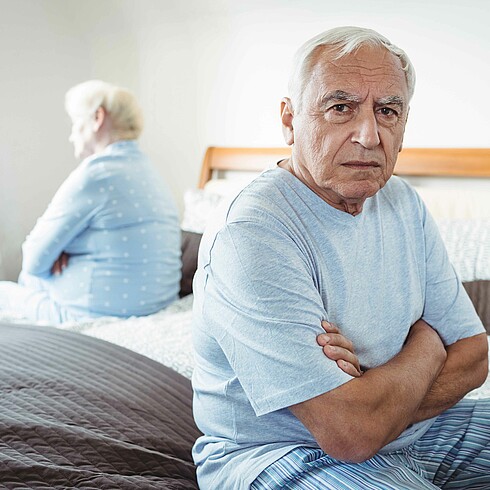 Mann sitzt besorgt auf dem Bett, im Hintergrund seine Frau