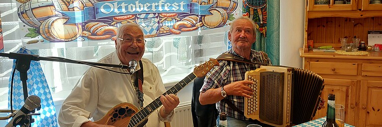Zwei ältere Männer musizieren mit Gitarre und Ziehharmonika vor Oktoberfest-Deko