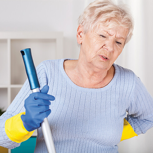 Ältere Dame mit Putzuntensilien hält sich den Rücken vor Schmerz