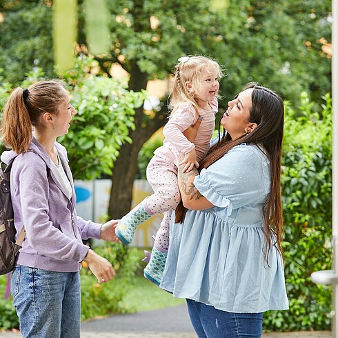 Mutter übergibt ihre Tochter den Armen der Kindergartenpädagogin, alle lachen fröhlich 