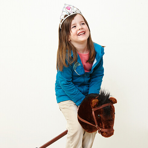 Mädchen mit Krone reitet auf einem Steckenpferd.