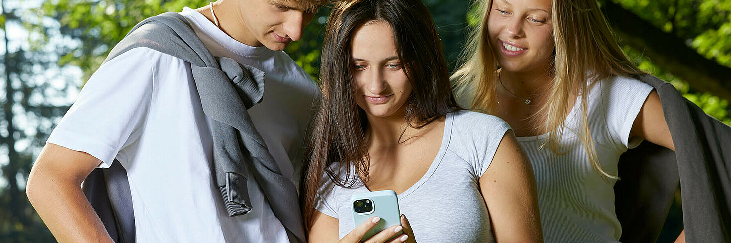 Drei Jugendliche in einem Park werfen einen gemeinsamen Blick auf ein Handy 