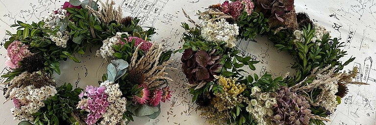Zwei Kränze aus grünen Zweigen, Trockenblumen und Gräsern liegen auf einer gezeichneten Karte.