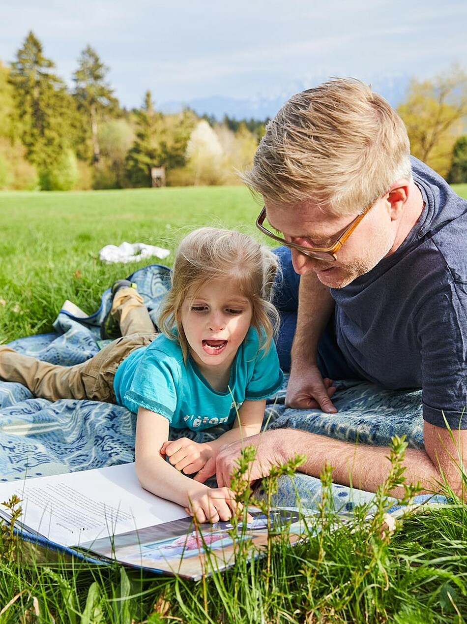 Vater und Tochter liegen auf Wiese und betrachten ein Bilderbuch