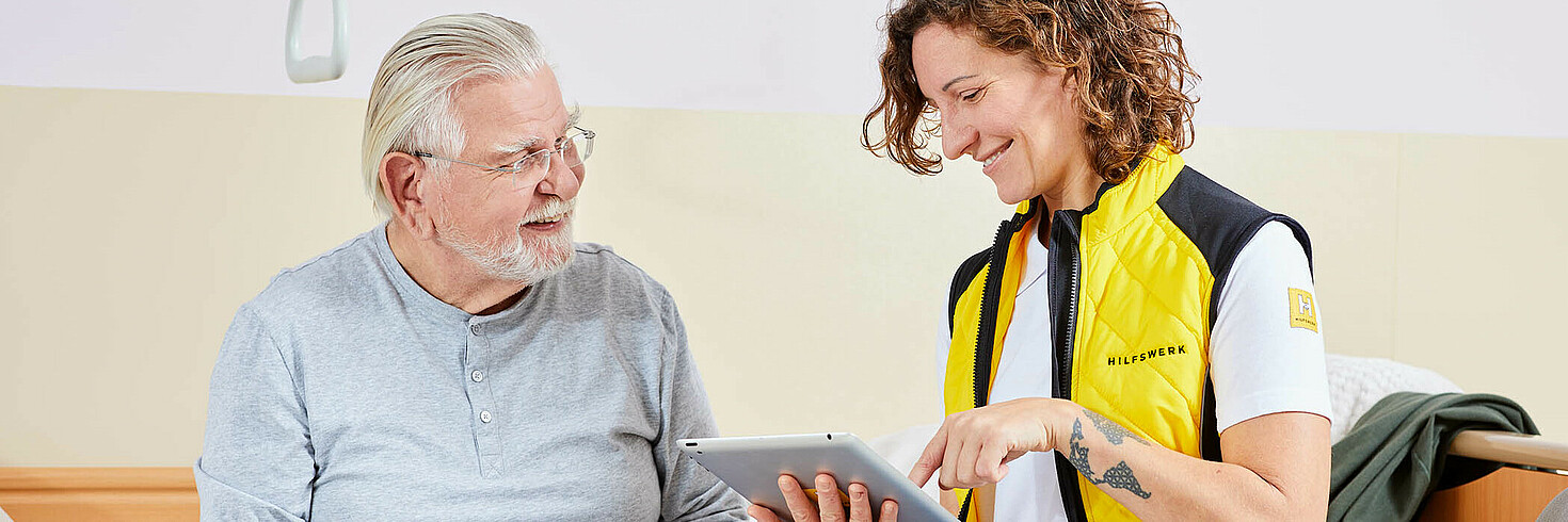 Ein älterer Herr und eine Hilfswerk Mitarbeiterin mit Tablet sitzen nebeneinander.