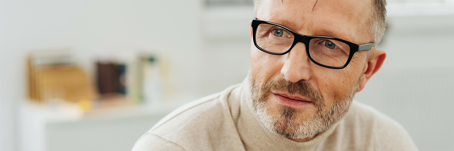Mann mittleren Alters mit Rollkragenpullover und Hornbrille in Nahaufnahme, er schaut links an der Kamera vorbei mit sinnierendem Blick 