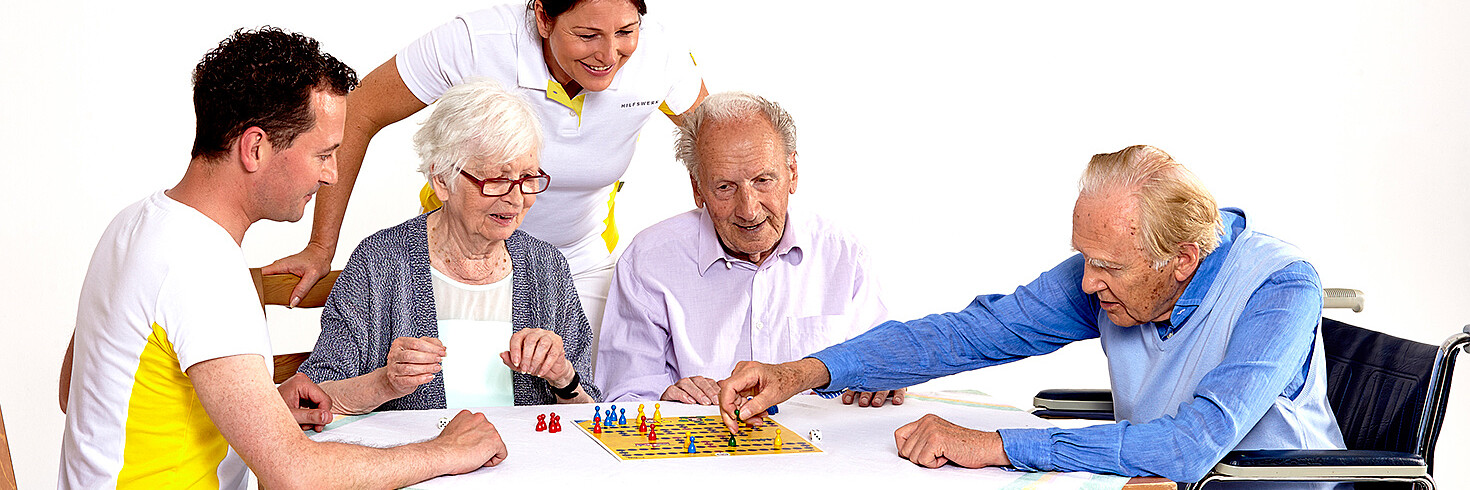 3 Seniorinnen und ein Senior, einer davon im rollstuhl, sitzen um einen Tisch und unterhalten sich, 2 Betreuer sind dahinter.