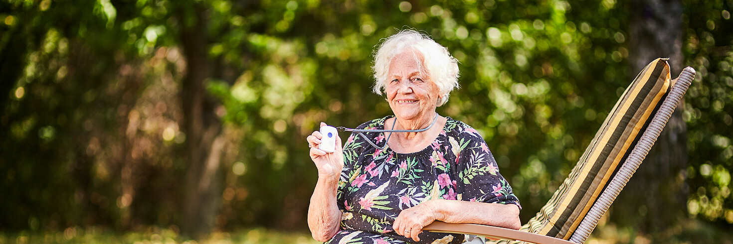 Ältere Dame mit Notruftelefon sitzt im Garten auf einer Liege.