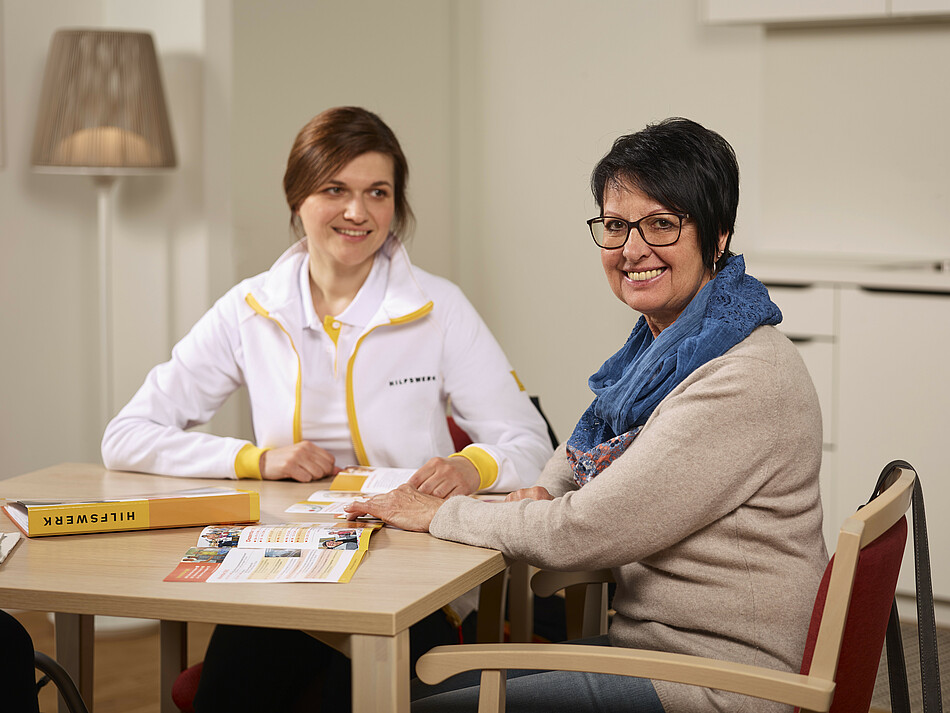 Hilfswerk-Mitarbeiterin sitzt mit einer Dame redend an einem Tisch.