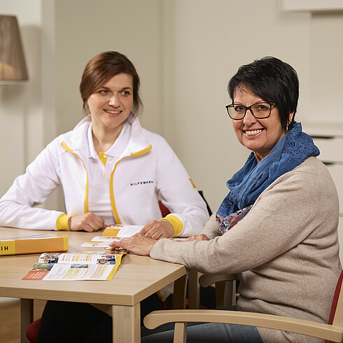 Hilfswerk-Mitarbeiterin sitzt mit einer Dame redend an einem Tisch.