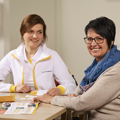 Hilfswerk-Mitarbeiterin sitzt mit einer Dame redend an einem Tisch.