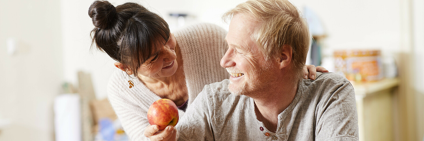 Eine Frau mittleren Alters beugt sich freundlich lächelnd von hinten über die Schulter ihres Partners und bietet ihm einen Apfel an.