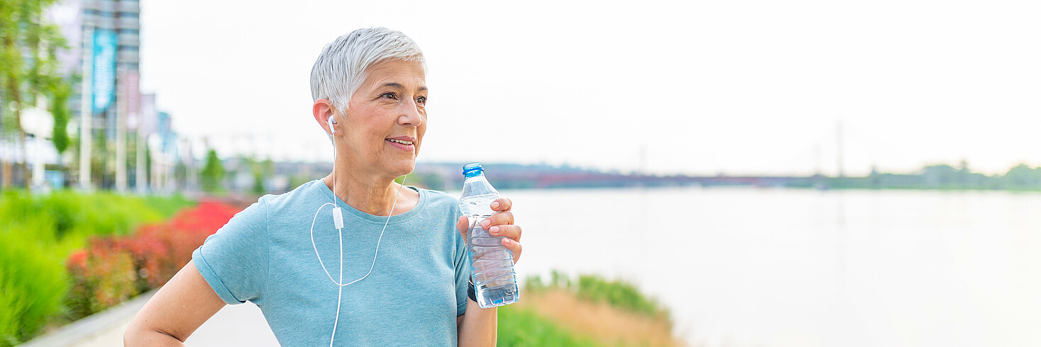 Eine Joggerin mittleren Alters steht am Fluss und hält eine Wasserflasche in der Hand. 