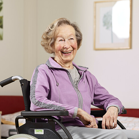 Ältere Dame sitzt lachend in einem Rollstuhl.