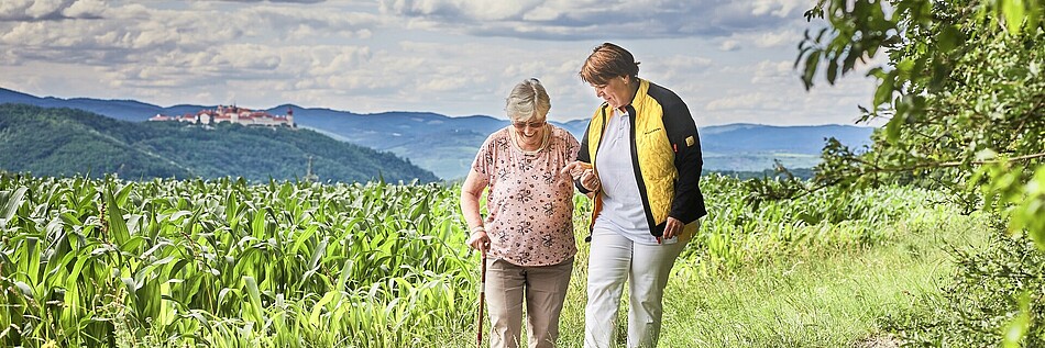 Eine Seniorin und eine Pflegerin spazieren über einen Feldweg