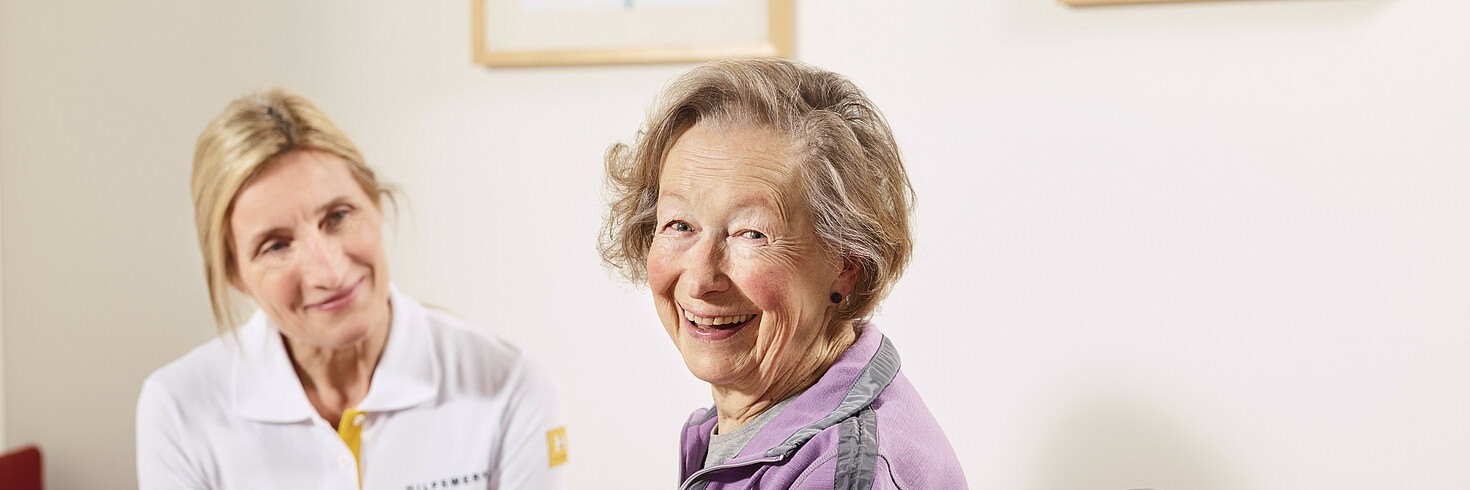 Hilfswerk-Mitarbeiterin hilft älterer Dame aus dem Bett in ihren Rollstuhl.