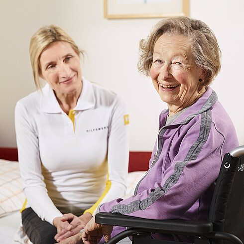 Hilfswerk-Mitarbeiterin hilft älterer Dame aus dem Bett in ihren Rollstuhl.
