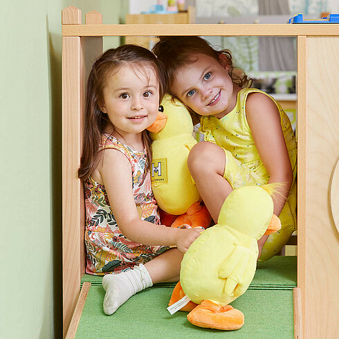 Zwei Mädchen mit gelber Plüschente auf in einem Indoor-Klettergestell, beide lächeln in die Kamera 