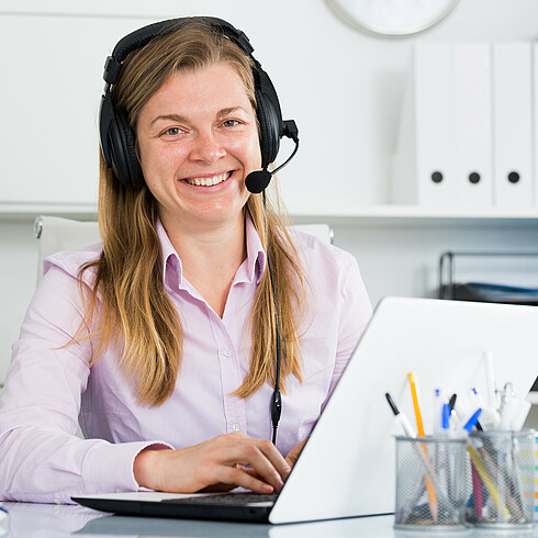 Junge Beraterin sitzt mit Headset vor dem Bildschirm und schaut lächelnd in die Kamera