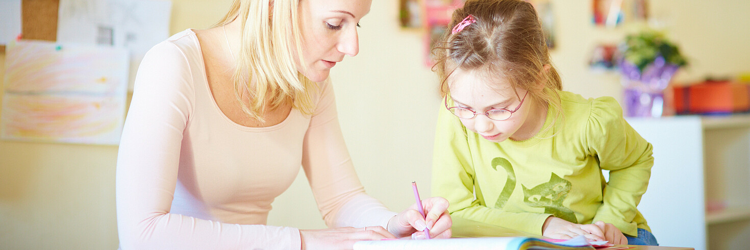 Ein junges Mädchen und eine Kinderbetreuerin sitzen an einem Tisch und machen Hausaufgaben.