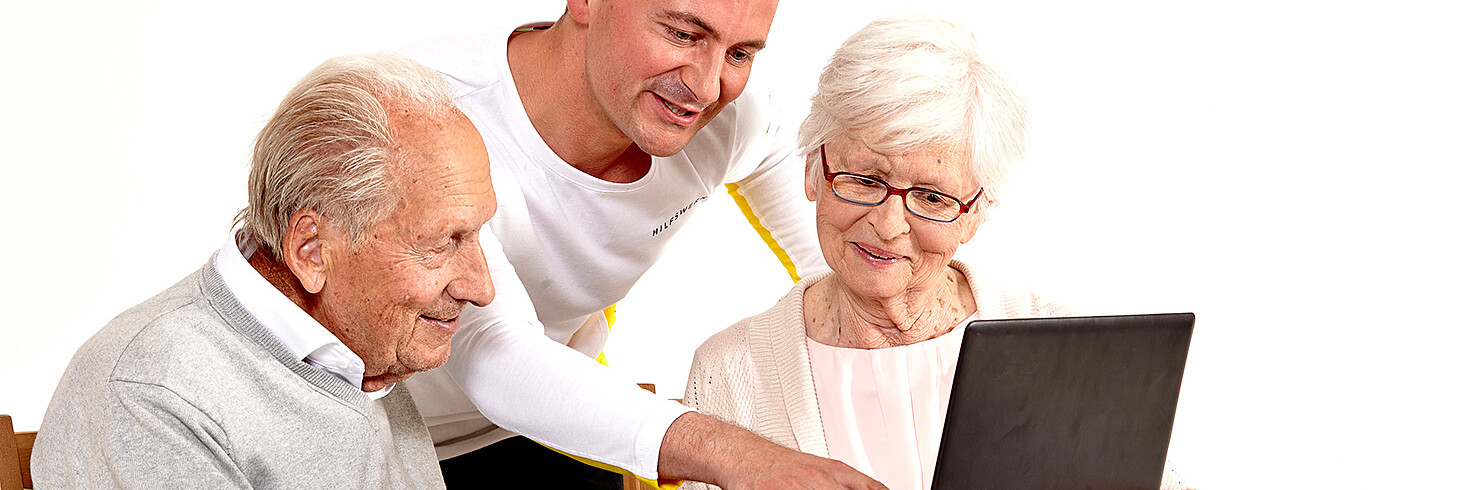 Ein Senior und eine Senioren sitzen vor einem Laptop. Der Betreuer erklärt etwas. 