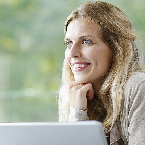 Junge Frau am Computer sitzend schaut lächelnd aus dem Fenster 