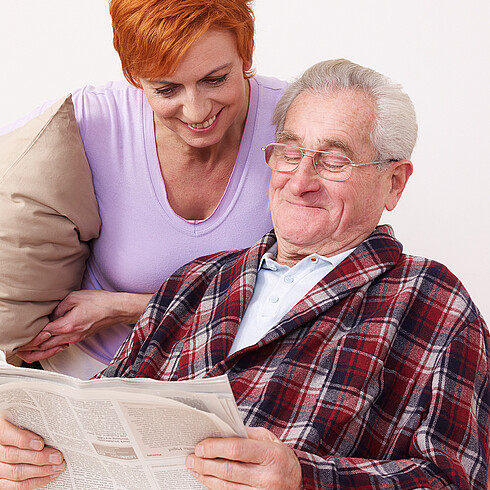 Betreuerin steht hinter älterem Herr der im Wohnzimmersessel sitzt und Zeitung liest 