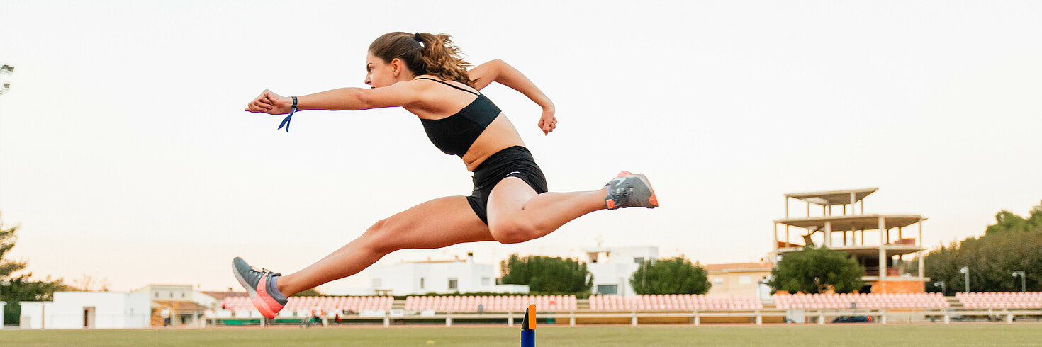 Eine junge Hürdenläuferin im Stadion springt von rechts nach links über eine Hürde 