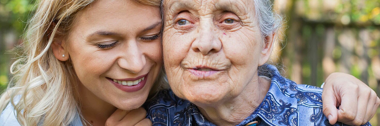 Jüngere Frau umarmt ältere Frau, ältere Frau schaut ins Leere, aber lächelt