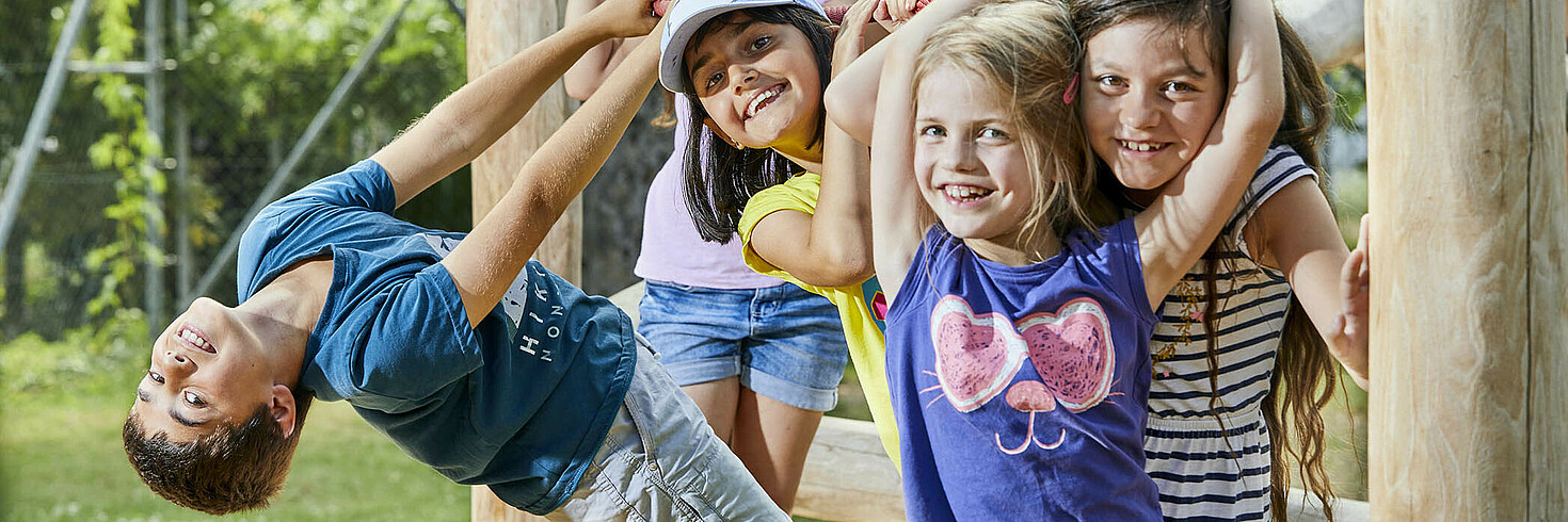 Gruppe von Kindern im Schulalter klammert sich ans Kletterseil eines Klettergestells, alle lachen in die Kamera
