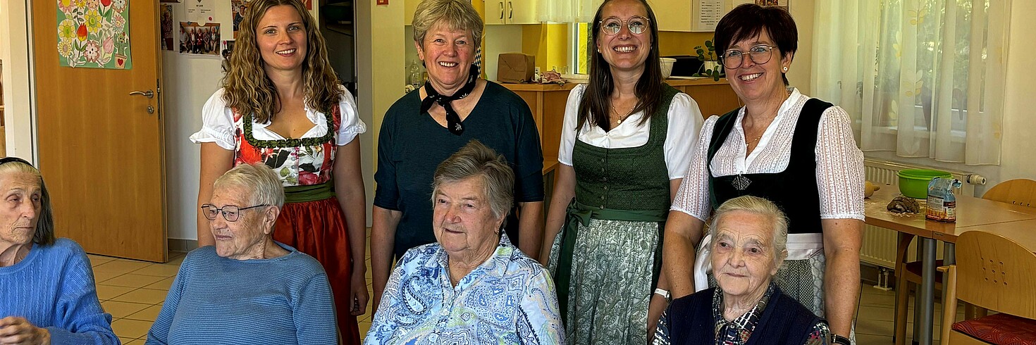 Drei Frauen in Tracht und zwei Mitarbeiterinnen lächeln in die Kamera, im Hintergrund sitzen zwei ältere Damen.