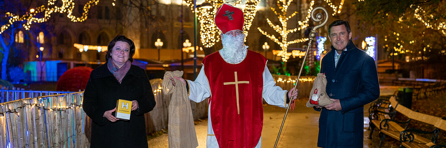 Der Nikolo eine Frau und ein Mann. Im Hintergrund weihnachtliche Beleuchtung.