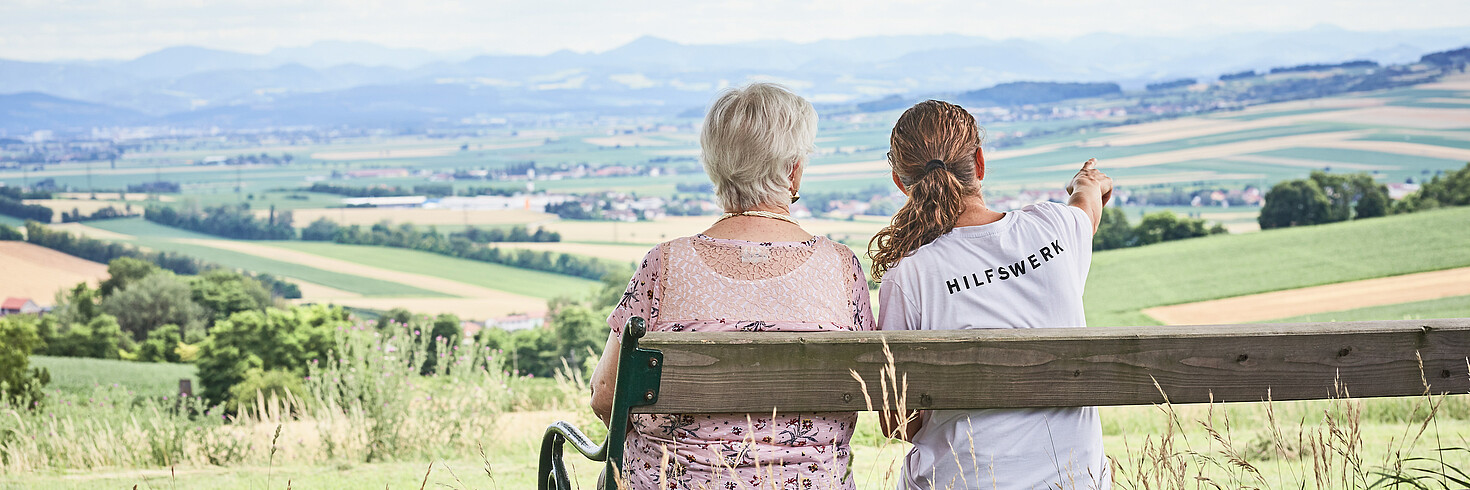 Hilfswerkerin sitzt mit einer älteren Dame auf einer Bank und blickt über die Landschaft