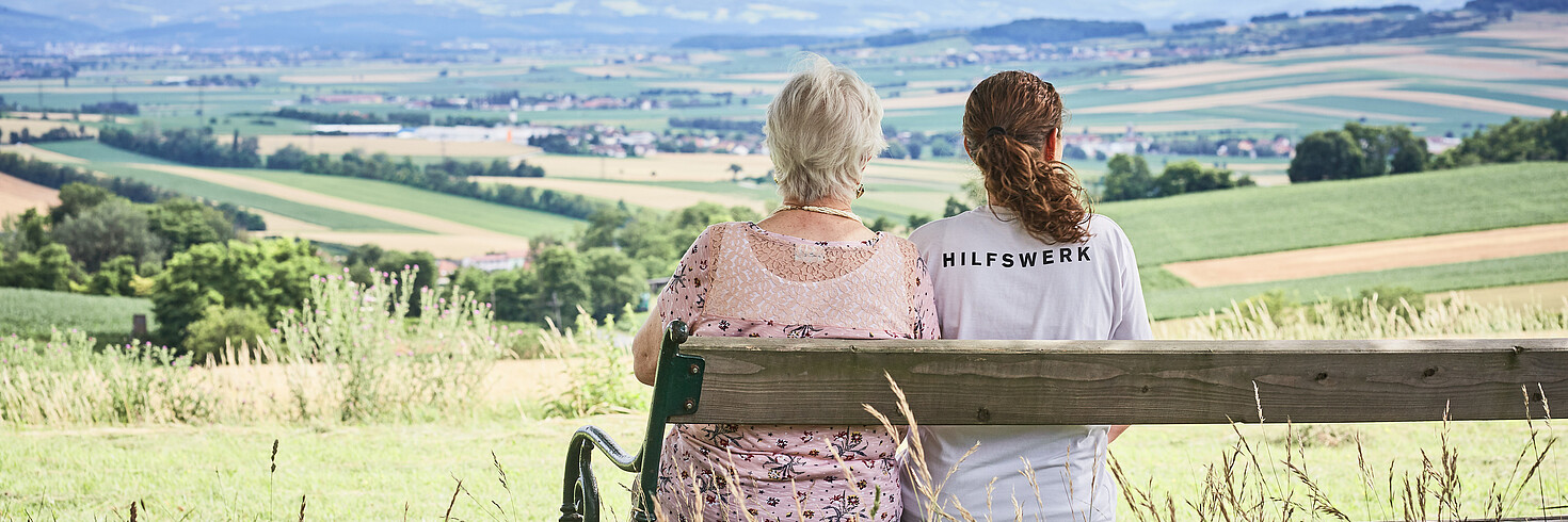 Hilfswerkerin sitzt mit einer älteren Dame auf einer Bank und blickt über die Landschaft