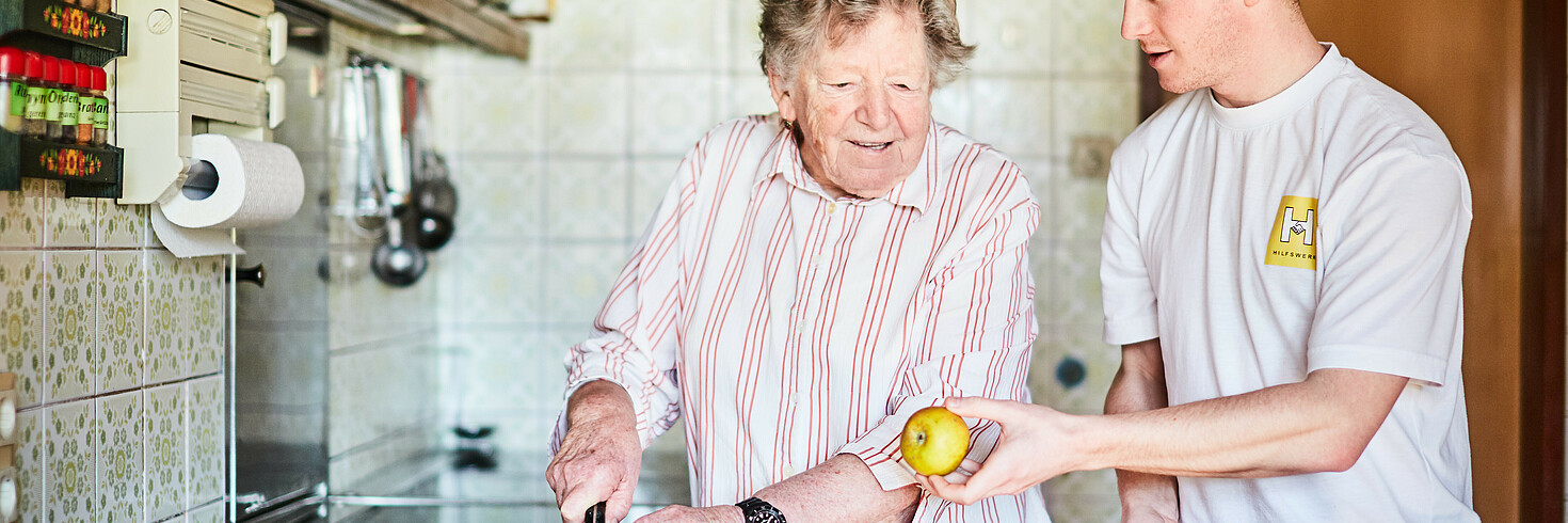 Heimhilfe unterstützt ältere Frau in ihrer Küche und gibt ihr Orangen